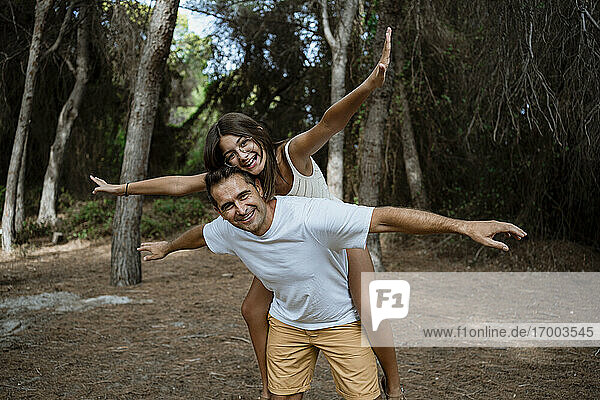 Smiling father giving piggyback ride to daughter with arms outstretched in forest during vacation