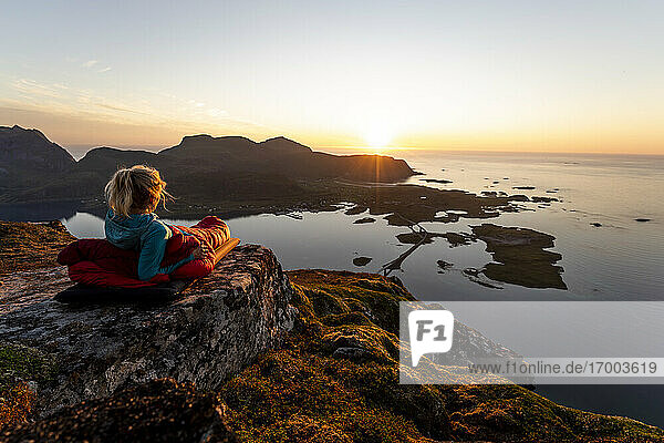 Wanderer bewundert die Aussicht auf den Sonnenuntergang  während er im Schlafsack auf dem Volandstinden  Lofoten  liegt