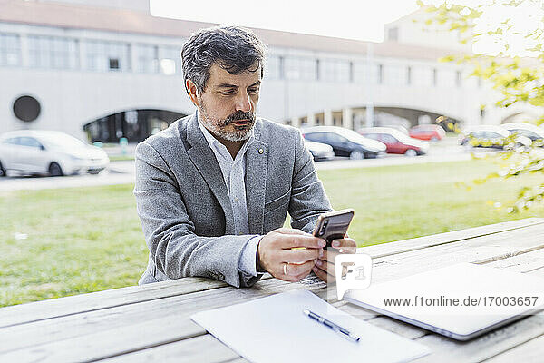 Reifer männlicher Berufstätiger  der ein Mobiltelefon am Tisch vor einem Bürogebäude benutzt