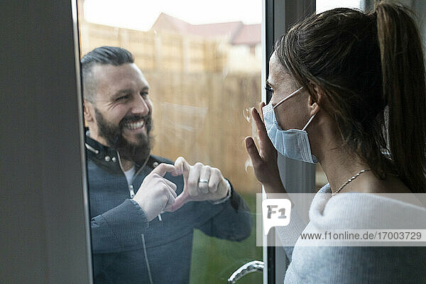 Freund zeigt seiner Freundin durch das Fensterglas eine herzförmige Geste während der COVID-19-Krise