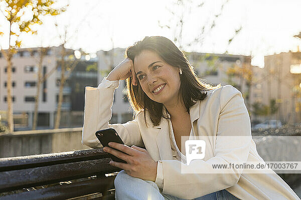 Nachdenklich lächelnde Geschäftsfrau mit Smartphone in der Hand auf einer Bank sitzend