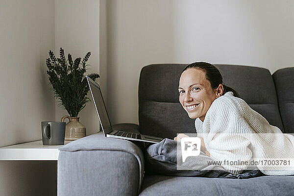 Smiling woman lying with laptop on sofa in living room