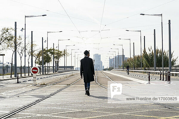 Modischer junger Mann  der zwischen Eisenbahnschienen auf einer Stadtstraße spazieren geht