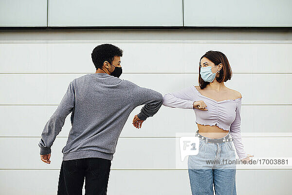 Junges Paar mit Gesichtsmaske grüßt mit Ellbogenstoß  während es an der Wand steht