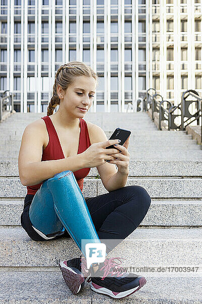 Sportlerin mit Beinprothese benutzt Mobiltelefon  während sie auf einer Treppe in der Stadt sitzt