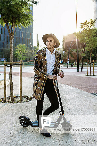 Stylish businessman in hat and checked jacket standing on push scooter in city