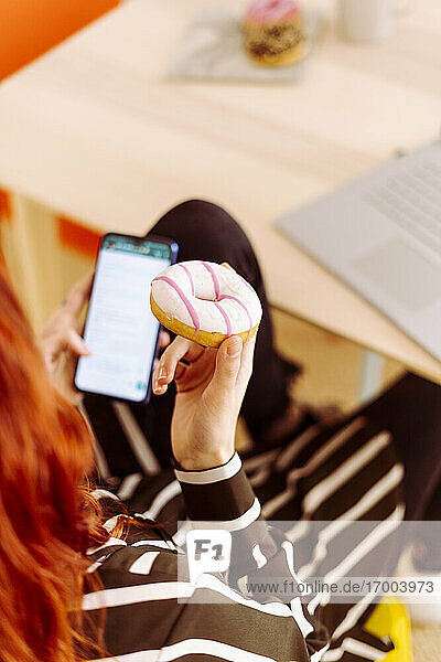 Geschäftsfrau  die einen Donut hält  während sie im Büro sitzt und ein Mobiltelefon benutzt