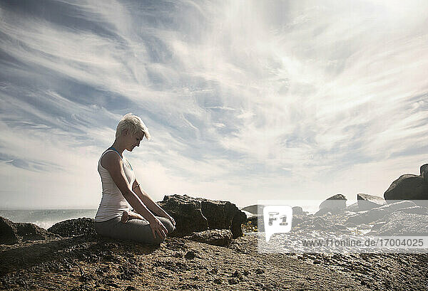 Frau meditiert bei der Ausübung von Yoga im Lotussitz auf einer Felsformation am Strand