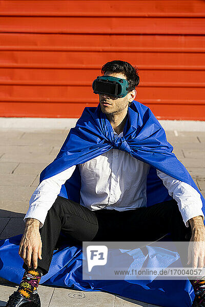 Geschäftsmann mit Superheldenumhang und VR-Brille sitzt auf dem Bordstein