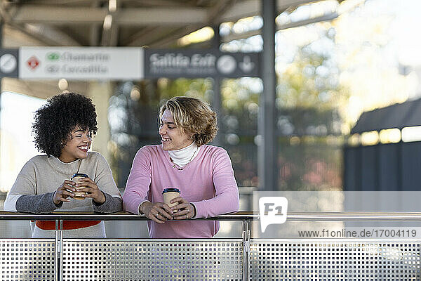 Fröhliche multiethnische Freunde halten wiederverwendbare Becher  während sie sich am Bahnhofsgeländer abstützen