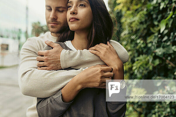 Junger romantischer Mann umarmt Frau  die wegschaut  während er im Park steht