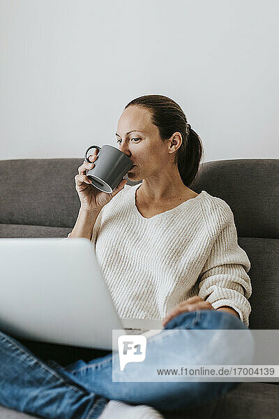 Frau trinkt Kaffee und benutzt einen Laptop zu Hause