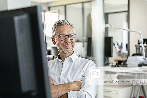 Smiling male professional with arms crossed day dreaming at work place