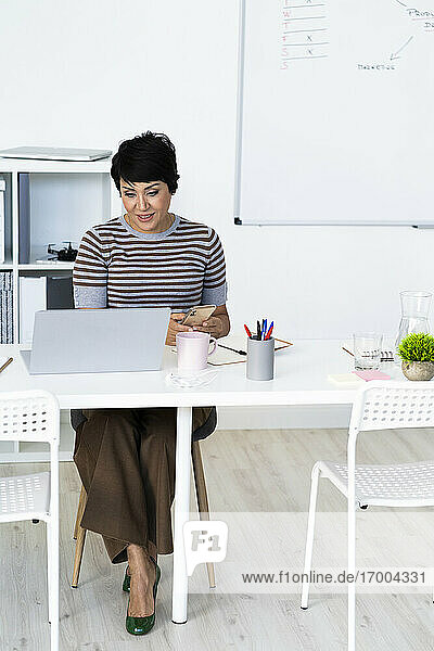 Porträt einer Geschäftsfrau  die am Bürotisch sitzt und ihr Smartphone in der Hand hält