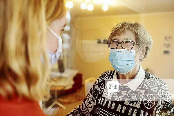 Großmutter mit Gesichtsmaske  die ihre Enkelin ansieht  während sie bei Covid-19 zu Hause am Glas steht