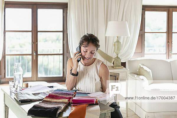 Geschäftsfrau  die mit ihrem Smartphone telefoniert  während sie zu Hause Farben aus Stoffmustern auswählt