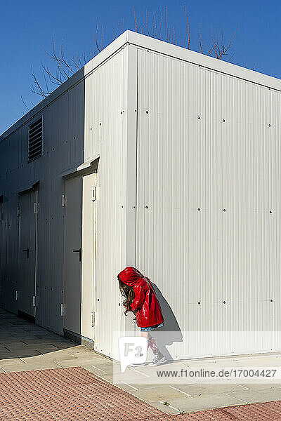 Little girl wearing red rain coat hiding behind a corner