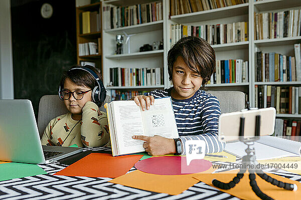 Junge zwinkert  während er bei einem Videoanruf mit einem männlichen Freund im Wohnzimmer sitzt und auf ein Lehrbuch zeigt