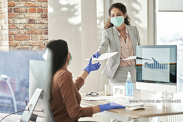 Young businesswoman giving report to female coworker while working in office during pandemic