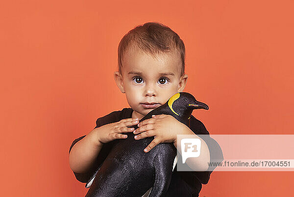 Süßes Baby Mädchen umarmt Pinguin Spielzeug  während gegen orange Hintergrund sitzen