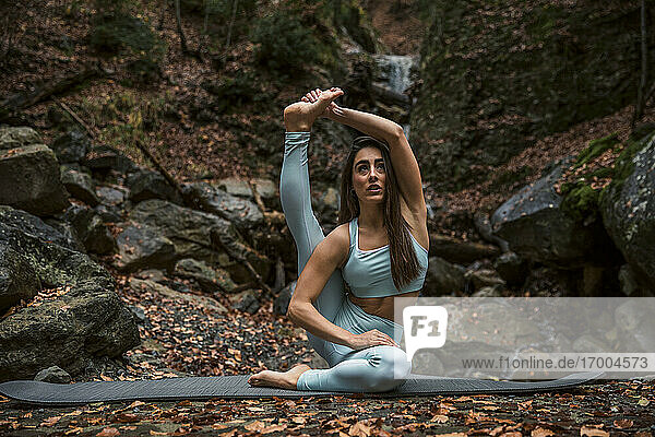 Athlete doing yoga while sitting on exercise mat against waterfall in forest
