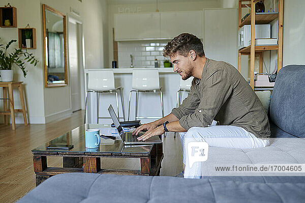 lächelnder männlicher Unternehmer  der zu Hause mit einem Laptop arbeitet  während er auf dem Sofa im Wohnzimmer sitzt