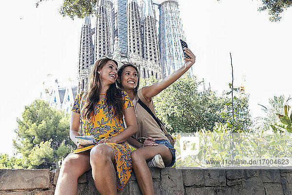 Lächelnde Freunde machen ein Selfie  während sie auf einer Stützmauer im Sagrada Familia Park sitzen  Barcelona  Katalonien  Spanien