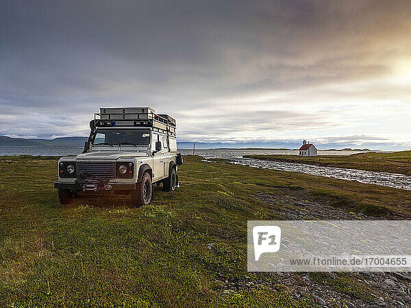 Off-road car parked in front of Icelandic river at cloudy sunset with secluded Unadsdalskirkja church in background