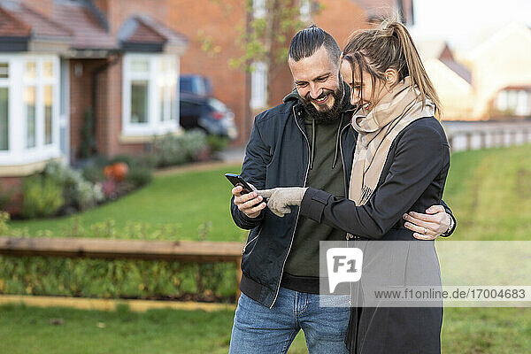 Glückliches Paar schaut auf sein Smartphone  während es im Vorgarten steht