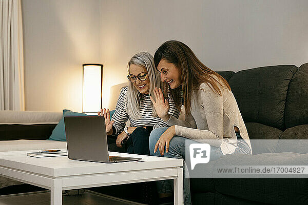 Lächelnde Frauen winken mit der Hand zum Videoanruf auf dem Laptop  während sie zu Hause auf dem Sofa sitzen