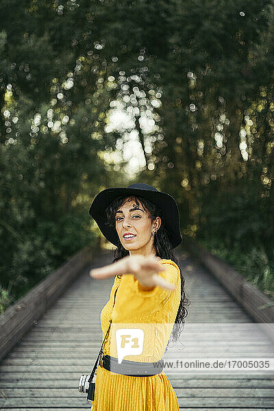 Junge Frau mit schwarzem Hut und gelbem Kleid mit einer analogen Kamera schüttelt die Hand zu einer Kamera auf der Holzpromenade