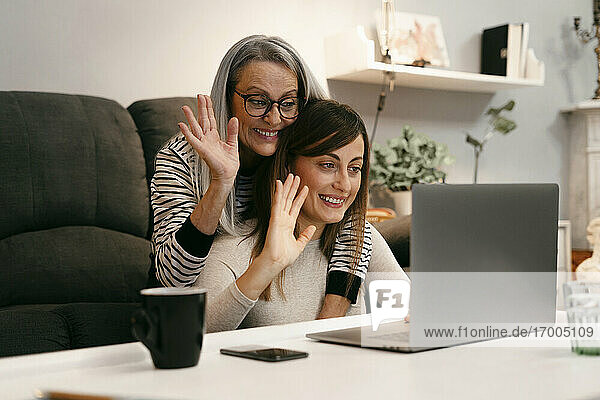 Glückliche Frauen winken mit der Hand zum Videoanruf auf dem Laptop  während sie zu Hause sitzen