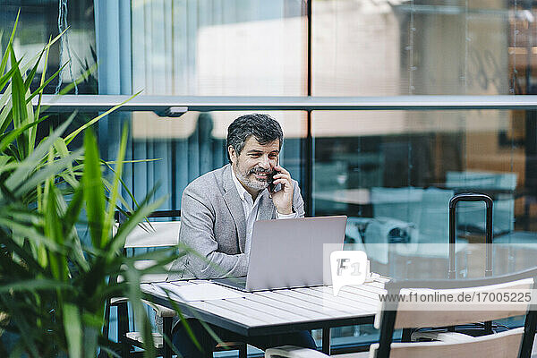 Lächelnder Geschäftsmann am Telefon mit Blick auf den Laptop in einem Cafe