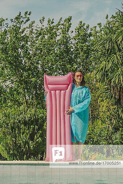 Woman in blue rain coat standing at poolside with pink airbed