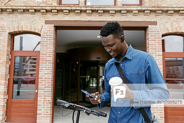 Lächelnder Geschäftsmann  der sein Smartphone benutzt und einen wiederverwendbaren Kaffeebecher vor einem Gebäudeeingang in der Stadt hält