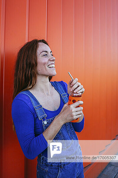 Lächelnde Frau  die einen Saft trinkt  während sie an einer orangefarbenen Wand steht