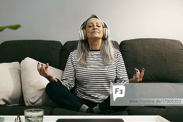 Lächelnde ältere Frau mit Kopfhörern  die zu Hause auf dem Sofa sitzt und meditiert