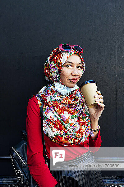 Lächelnde Frau hält Einweg-Kaffeebecher gegen schwarze Wand während COVID-19