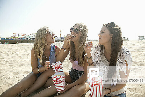 Smiling friends eating popcorn at beach on sunny day