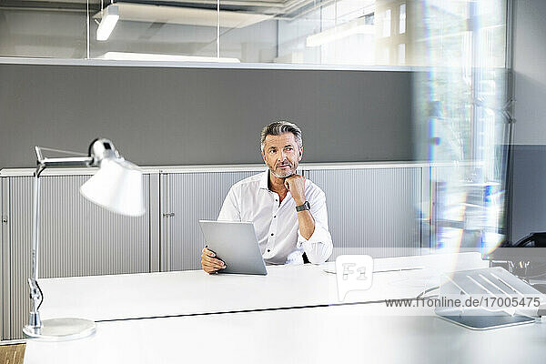 Nachdenklicher Geschäftsmann mit digitalem Tablet  der wegschaut  während er im Büro sitzt