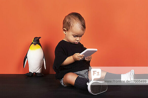 Baby-Mädchen spielen in Handy  während sie von Spielzeug gegen orange Hintergrund sitzen