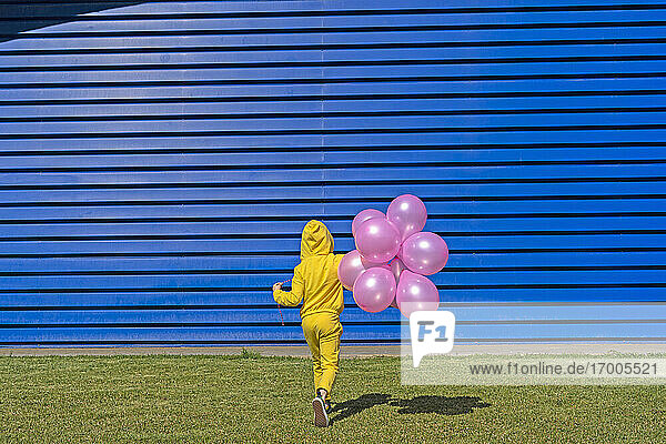 Back view of girl with pink balloons wearing yellow tracksuit walking in front of blue background