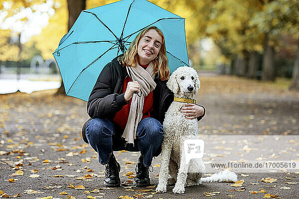 Smiling teenage girl holding umbrella while crouching by pet on road at park