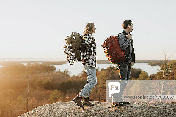 Junges Paar steht am Rande eines Felsvorsprungs und bewundert den umliegenden Wald bei Sonnenuntergang im Herbst