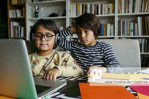 Junge schaut mit der Hand im Haar auf den Laptop  während er mit einem männlichen Freund im Wohnzimmer Hausunterricht erteilt