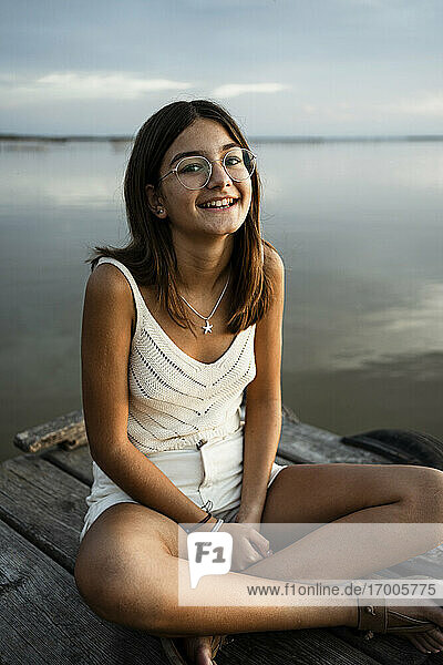 Mädchen mit Brille sitzt lächelnd auf einem Steg am See