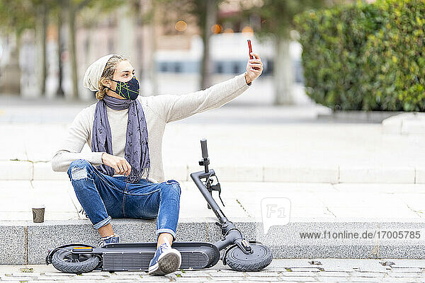 Mann mit Gesichtsmaske  der ein Selfie mit seinem Mobiltelefon macht  während er mit einem elektrischen Roller auf dem Gehweg sitzt