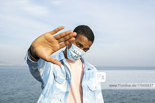Young man wearing Silence Is Violence written face mask showing stop gesture while standing against sea
