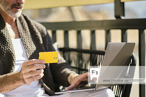 Älterer Mann zu Hause mit Kreditkarte beim Online-Shopping auf dem Laptop an einem sonnigen Tag