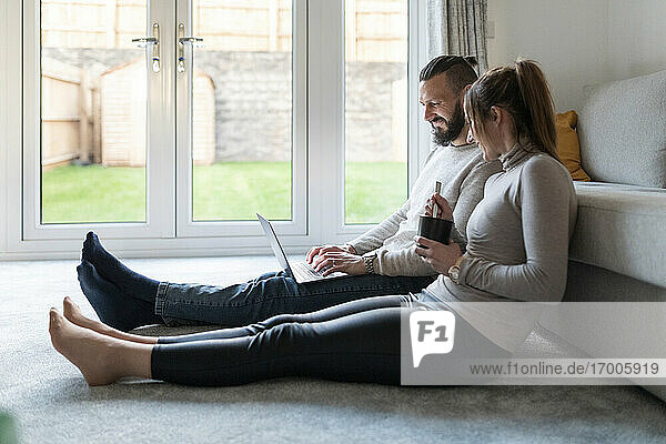 Lächelnde junge Frau trinkt Tee  während ihr Freund zu Hause einen Laptop benutzt